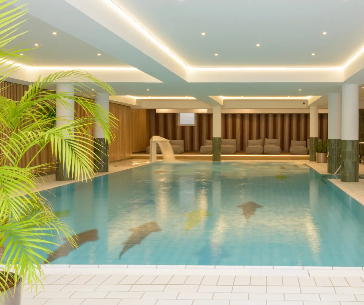 Das Hallenbad im Hotel Bodenmais besticht durch kristallklares Wasser, umgeben von eleganten Holzpaneelen und gemütlichen Liegestühlen. Eine üppige Pflanze im Vordergrund unterstreicht die ruhige Atmosphäre.