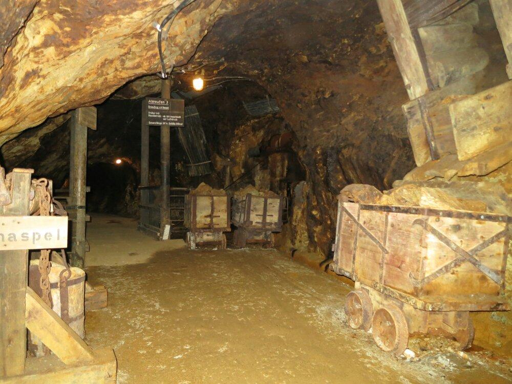 Unterirdischer Minentunnel mit hölzernen Stützbalken und verrosteten Erzkarren voller Steine.