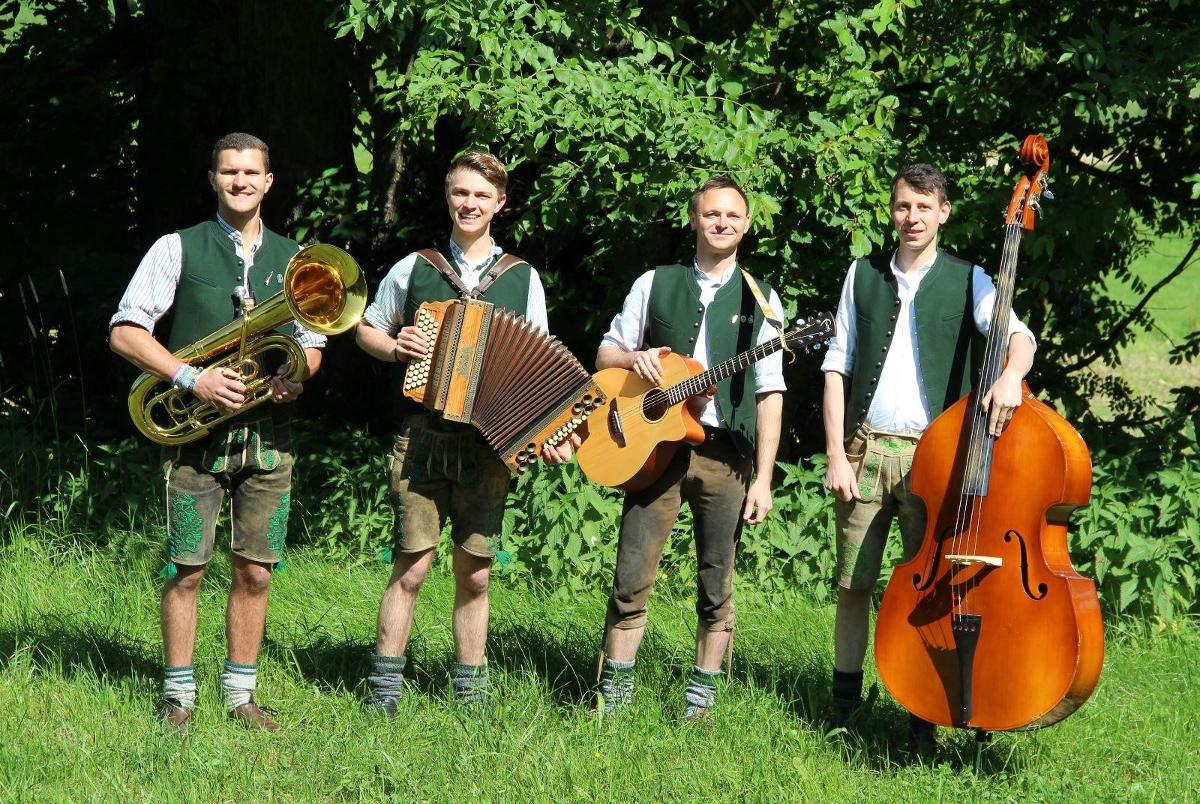 Vier Männer in traditioneller bayerischer Kleidung spielen im Freien im Sonnenlicht Blechbläser, Akkordeon, Gitarre und Kontrabass.