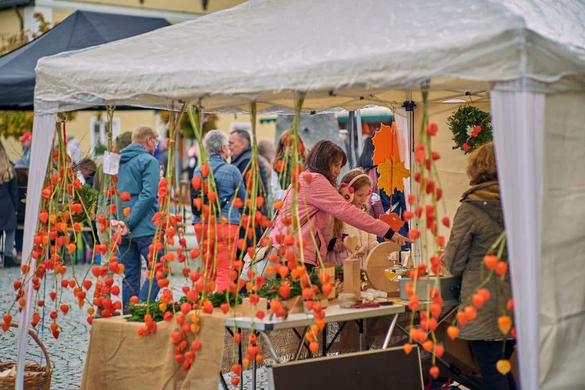 Menschen stöbern an einem Marktstand im Freien, der mit orangefarbenen Hängeornamenten und Herbstblättern geschmückt ist.