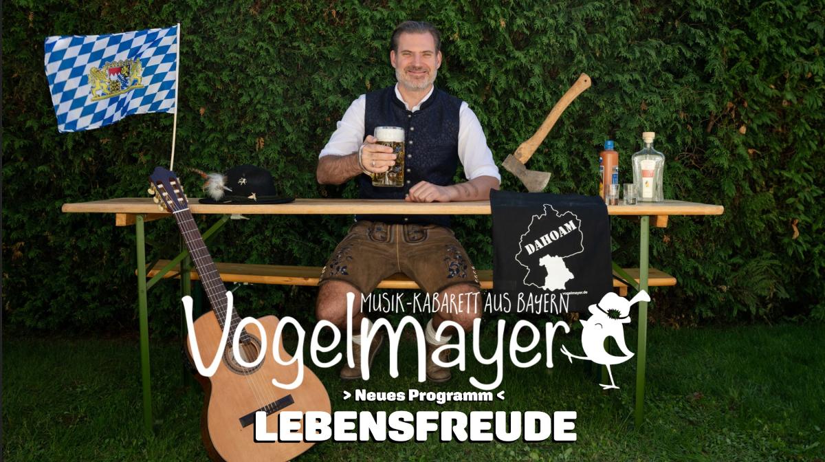 Mann in bayerischer Tracht sitzt an einem Tisch im Freien mit Bier, Gitarre und festlicher Dekoration. Text: Vogelmayer Lebensfreude.