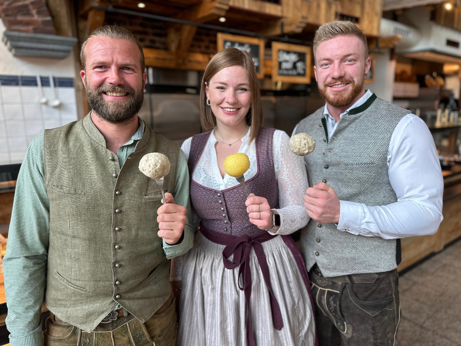 Drei lächelnde Menschen in traditioneller bayerischer Kleidung halten große Knödel auf Löffeln in einem rustikalen Restaurant.