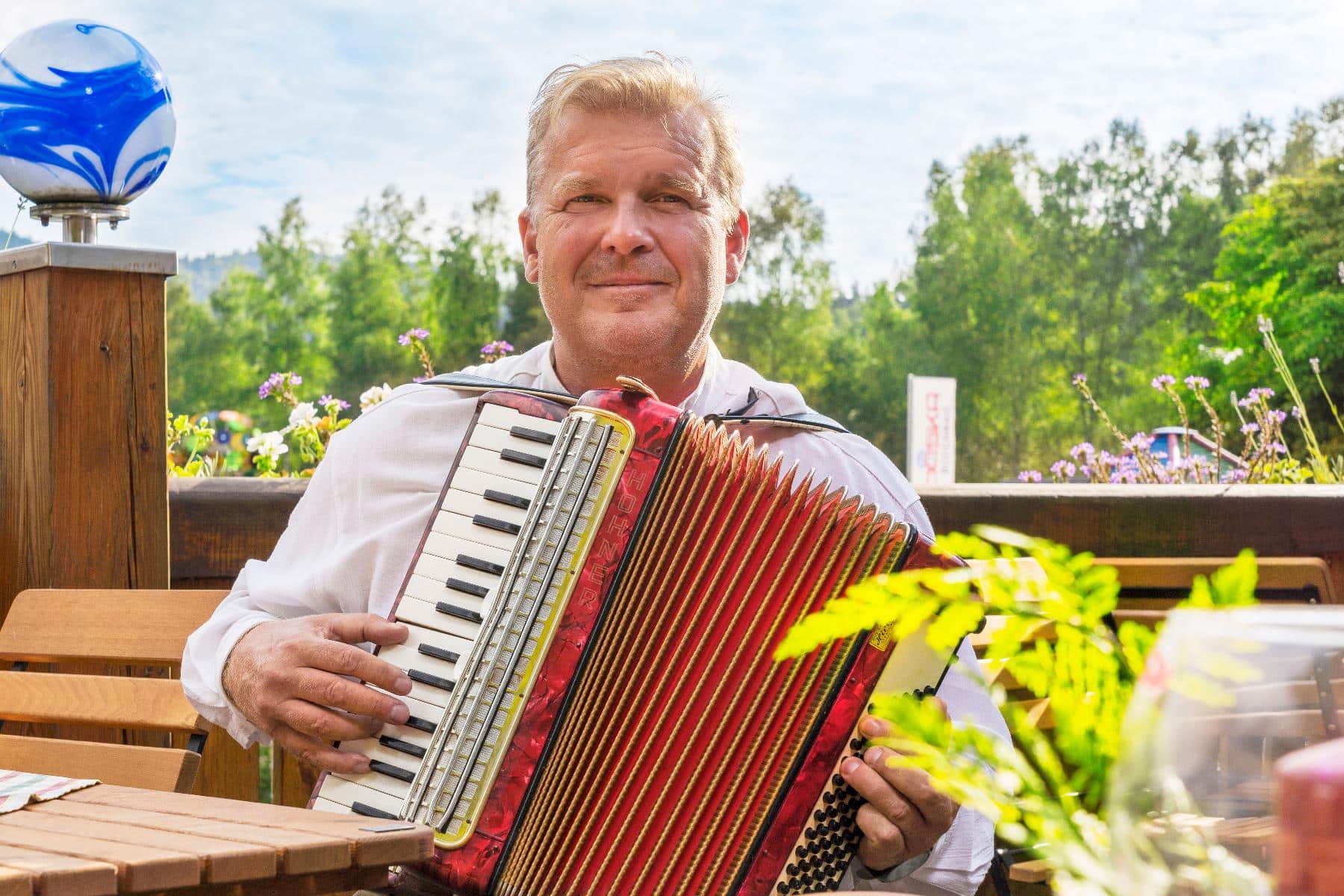 Mann mit blondem Haar spielt ein rotes Akkordeon im Freien, an einem Holztisch sitzend, an einem sonnigen Tag.