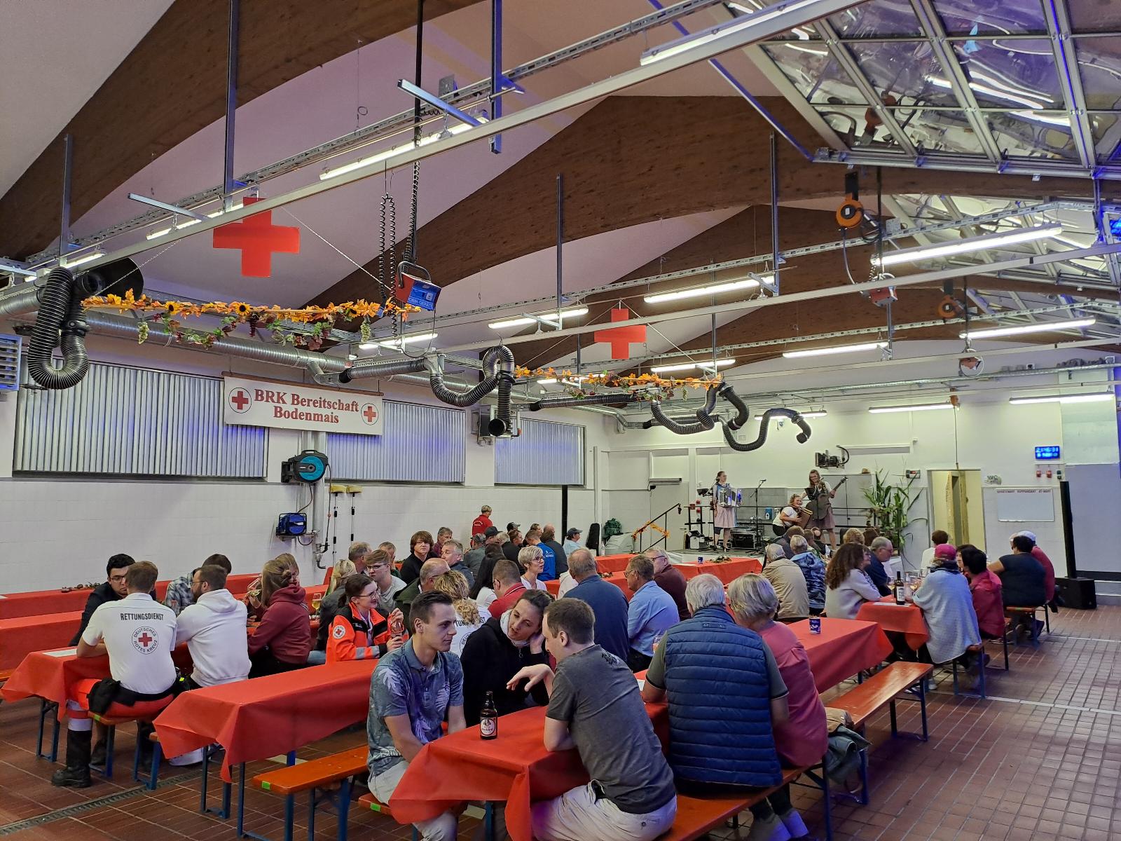 Die Menschen sitzen an roten Tischen in einer Halle mit Rot-Kreuz-Schildern und nehmen an einer Gemeinschaftsveranstaltung oder einem Treffen teil.