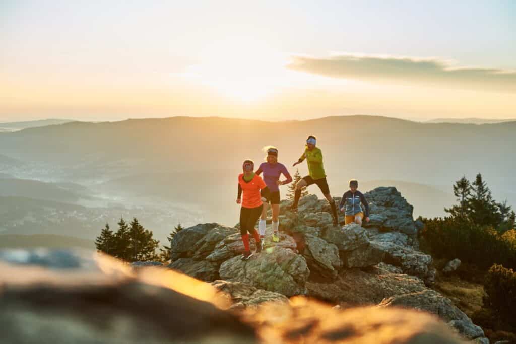Vier sportlich gekleidete Personen stehen bei Sonnenaufgang auf einem felsigen Gel&auml;nde und blicken auf neblige Berge und T&auml;ler.
