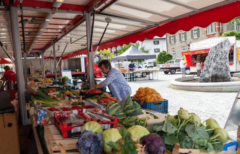 Eine Frau arrangiert Gemüse an einem Marktstand im Freien mit bunten Produkten und einem Stadtplatz im Hintergrund.