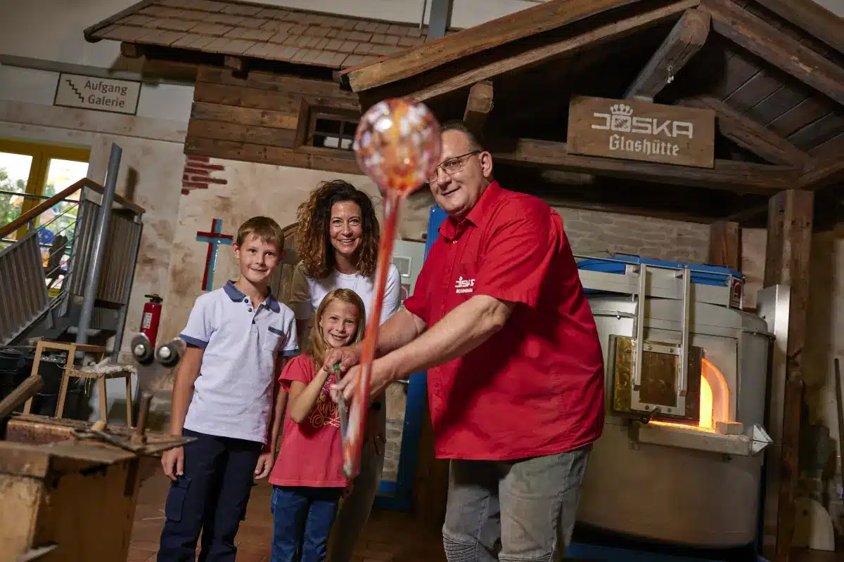 Ein Glasbläser führt einer lächelnden Familie in einer Glasbläserwerkstatt etwas vor.