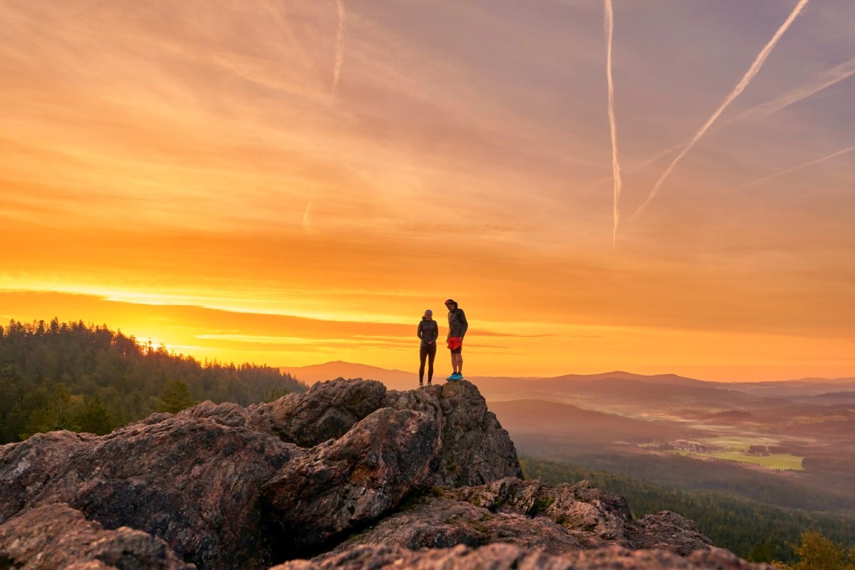 Der Bayerische Wald ist ein echter Geheimtipp: Zwei Wanderer stehen bei Sonnenaufgang auf einem felsigen Gipfel und blicken auf eine malerische Landschaft mit einem leuchtend orangefarbenen Himmel.