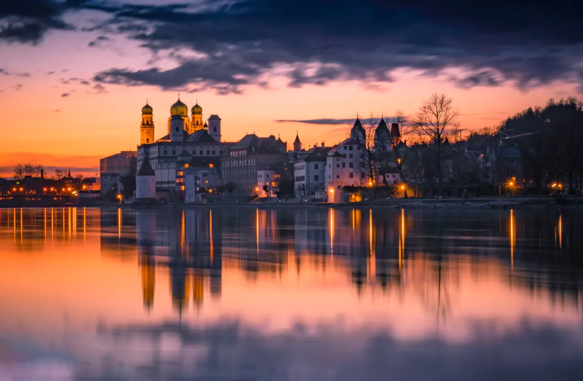 Stadtsilhouette der Stadt Passau mit Kirchtürmen, die sich im ruhigen Fluss bei Sonnenuntergang unter einem dramatischen, farbenfrohen Himmel spiegeln.