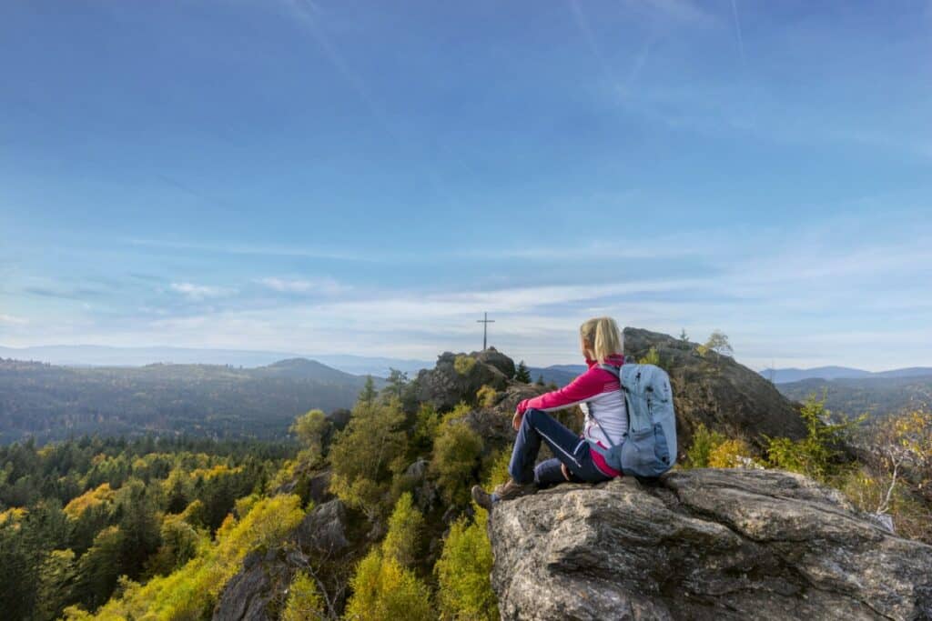 Eine Frau mit einem Rucksack sitzt auf einem Felsen und blickt auf einen Wald und ferne Berge unter blauem Himmel.