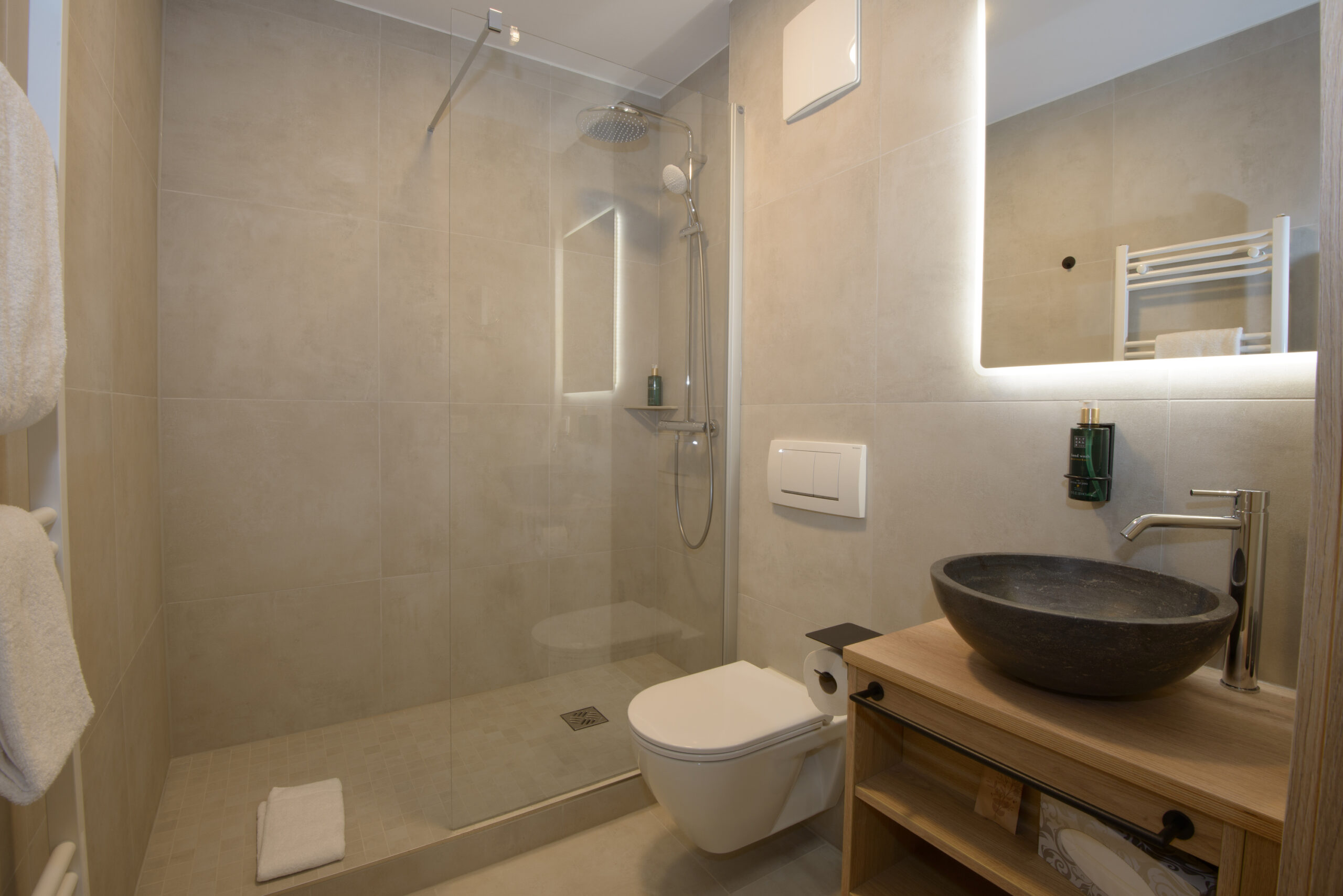 Modernes Badezimmer mit begehbarer Dusche, Waschbecken, Wandtoilette und großem beleuchtetem Spiegel.