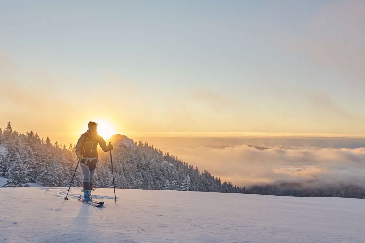 Eine Person beim Skifahren auf verschneitem Gelände bei Sonnenaufgang mit schneebedeckten Bäumen und Wolken im Hintergrund.