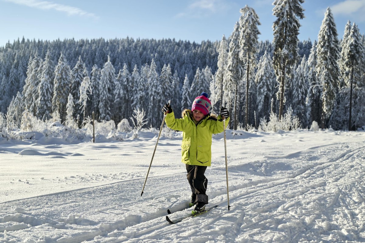 Ein Kind in einer hellen Jacke fährt Ski auf einer verschneiten Piste mit schneebedeckten Bäumen im Hintergrund.