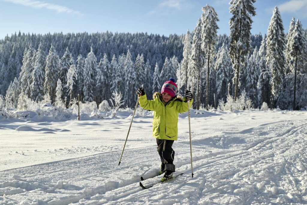 Kind in einer hellen Jacke beim Skilanglauf in Bodenmais auf einer verschneiten Loipe mit schneebedeckten B&auml;umen im Hintergrund.