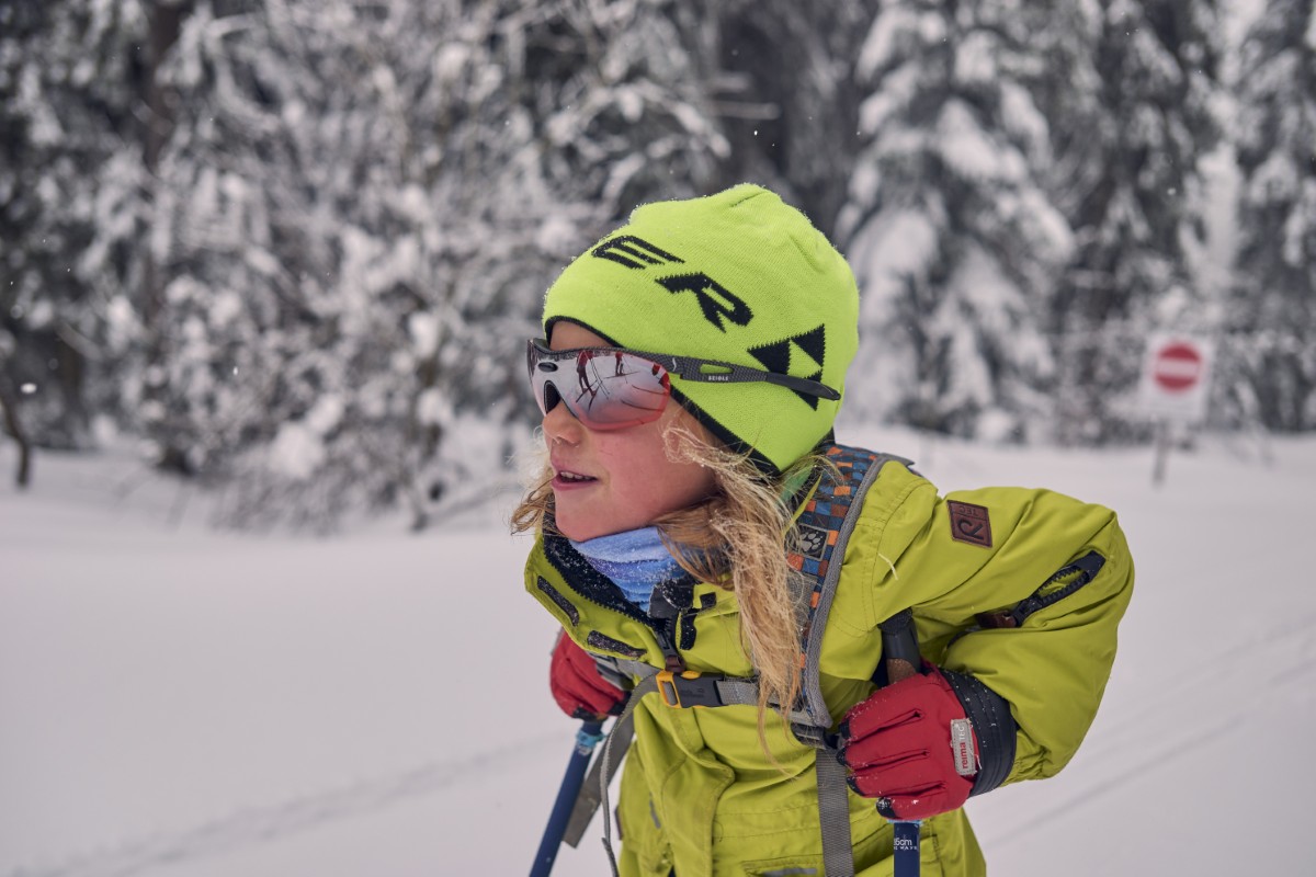 Ein Kind mit grüner Jacke und Mütze fährt auf Skiern durch einen verschneiten Wald, trägt eine Sonnenbrille und hat Skistöcke dabei.