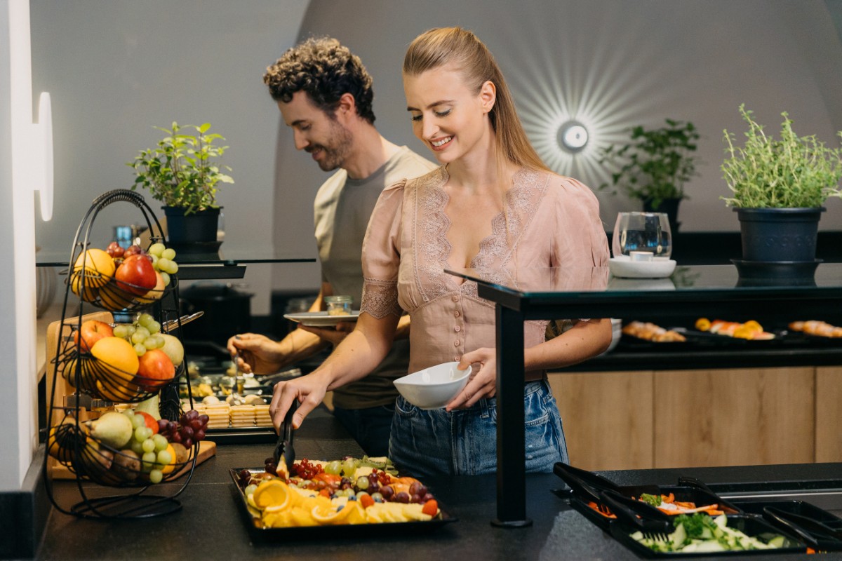 Eine Frau bedient sich an einem Buffet mit Obst, während ein Mann im Hintergrund Lebensmittel auswählt.