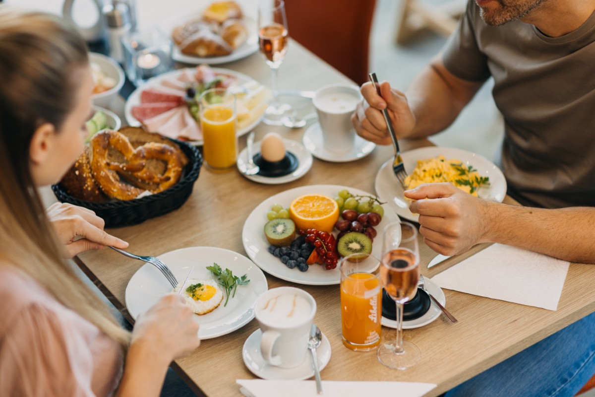 Zwei Personen essen an einem Tisch ein Frühstück mit Obst, Eiern, Brezeln, Saft und Kaffee.