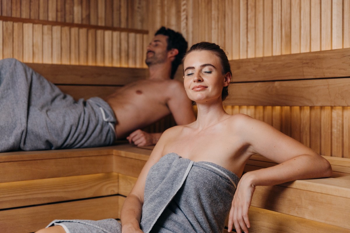 Zwei Personen, die sich in einer Holzsauna entspannen, beide in Handtücher gehüllt, mit heiterem Gesichtsausdruck.