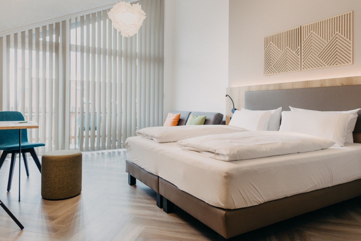 Modernes Hotelzimmer mit einem großen Bett, Kissen, einem Tisch mit Stühlen und vertikalen Jalousien vor den hohen Fenstern.