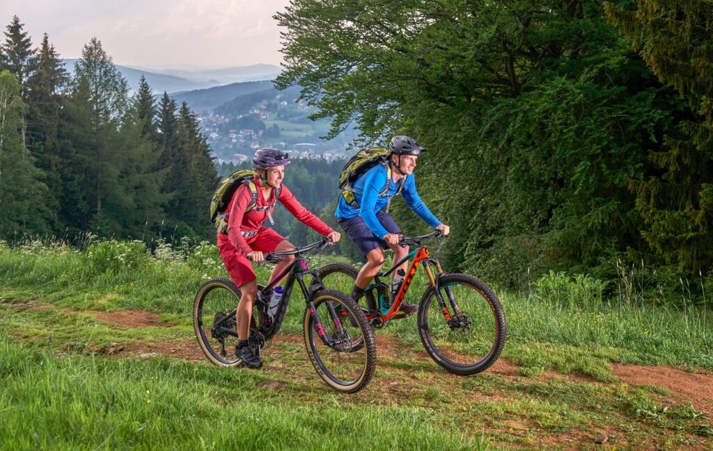 Zwei Personen fahren mit dem Mountainbike auf einem grasbewachsenen Weg durch ein bewaldetes Gebiet mit Hügeln im Hintergrund und genießen einen abenteuerlichen Tag in der Nähe des Aktivhotels Bayern.