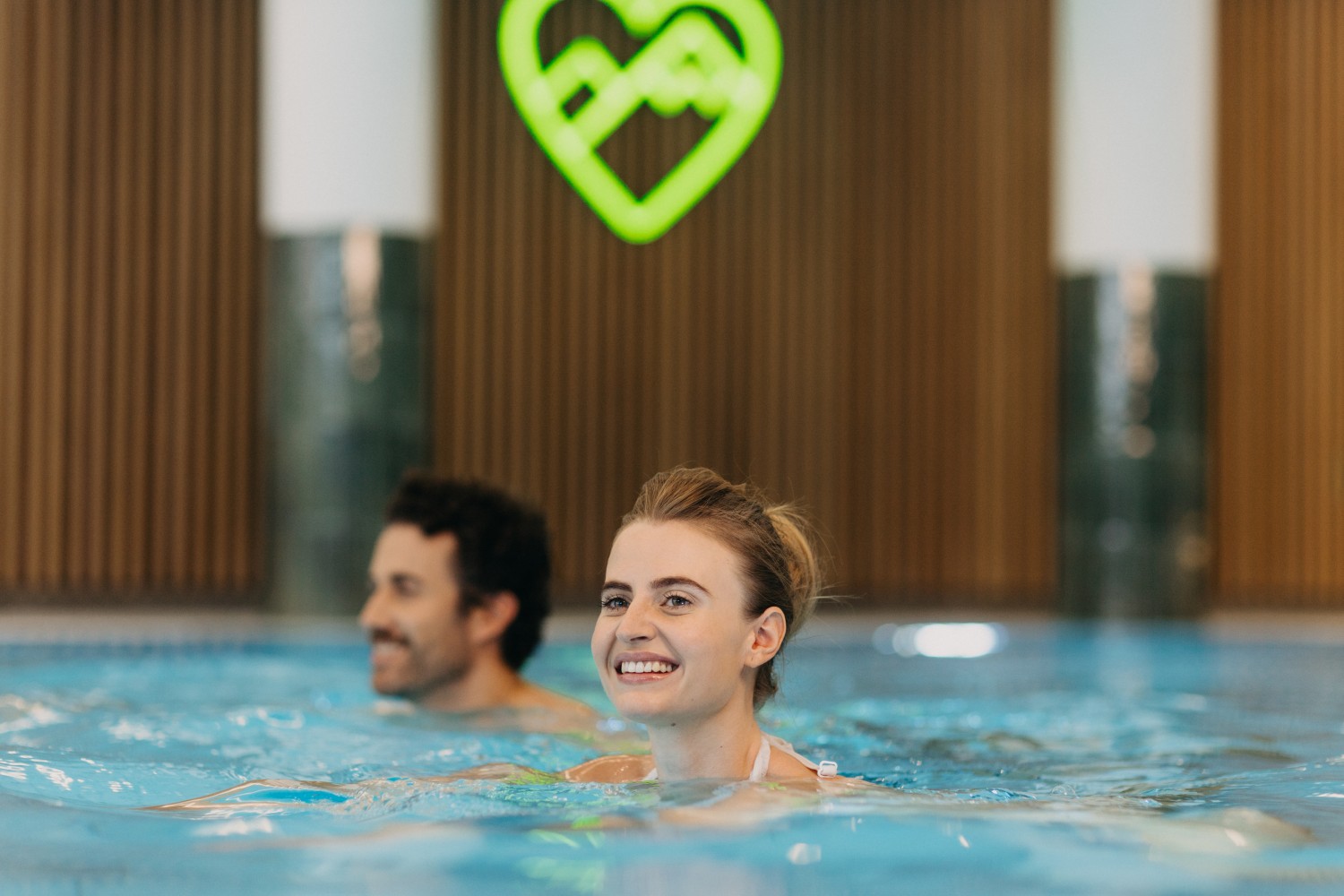 Lächelnde Frau und Mann beim Schwimmen im Hallenbad des Aktivhotels Bayern, mit einem leuchtenden herzförmigen Schild im Hintergrund.