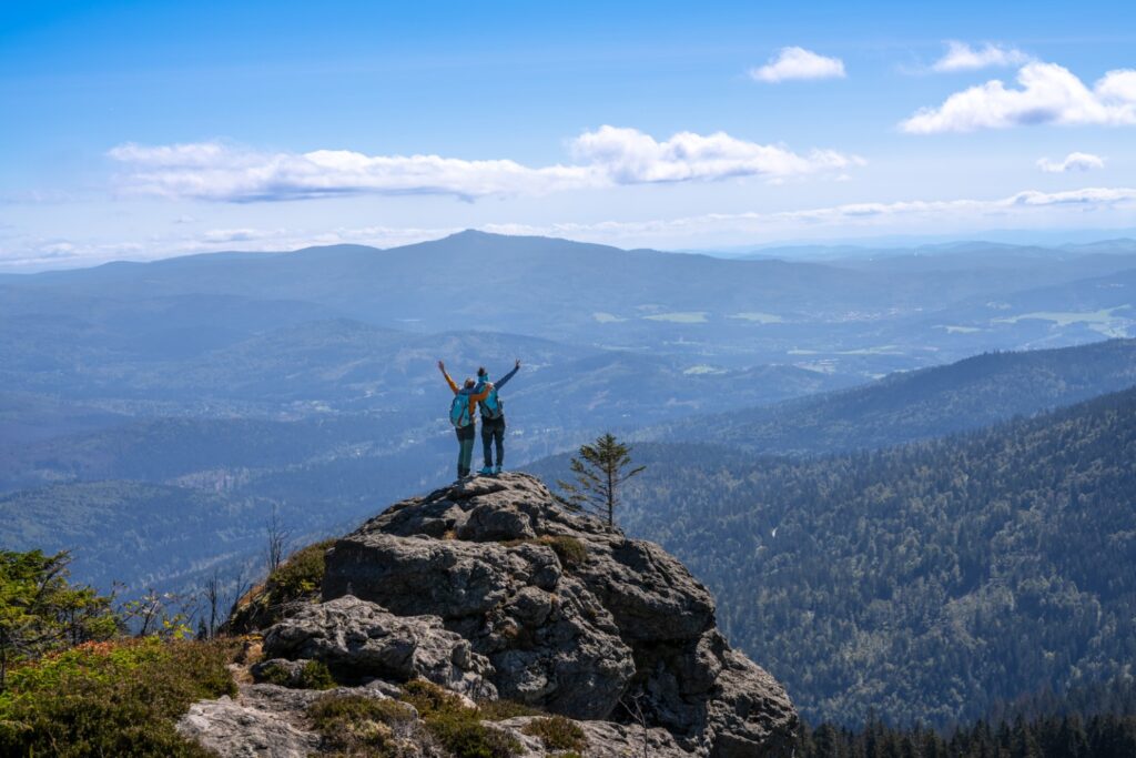 Zwei Wanderer stehen mit erhobenen Armen auf einem felsigen Gipfel und &uuml;berblicken eine weite Berglandschaft unter blauem Himmel - ein Abenteuer, inspiriert vom Geist des Aktivhotels Bayern.