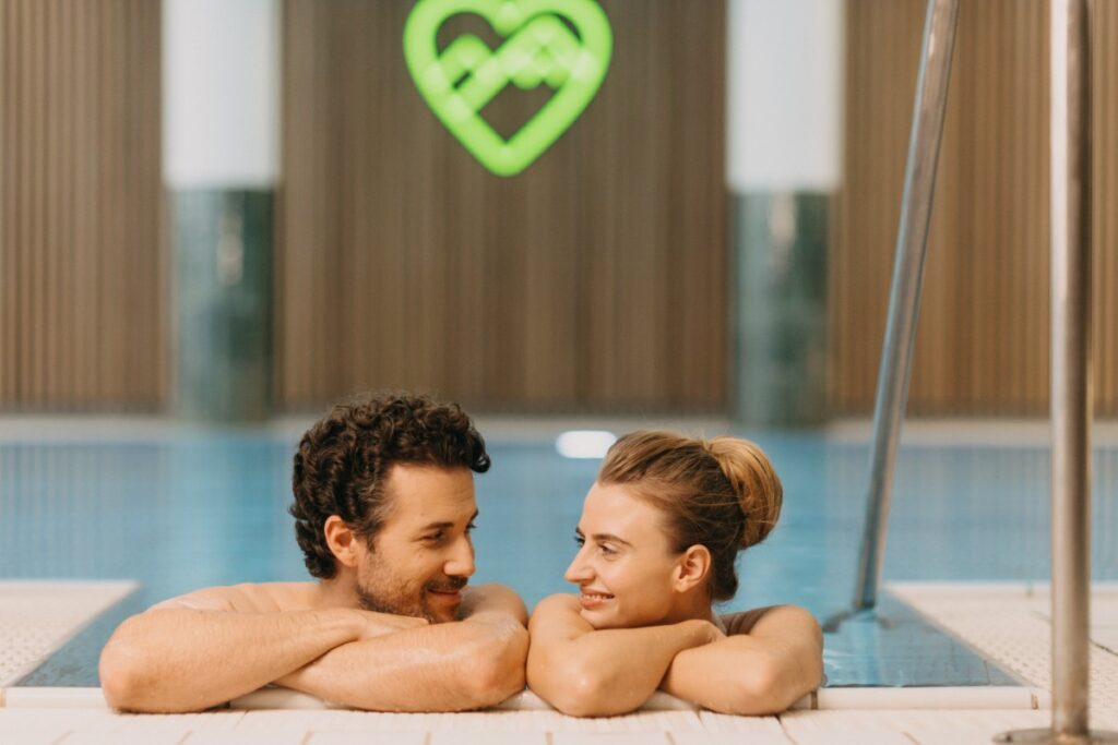 Ein Mann und eine Frau l&auml;cheln sich an, w&auml;hrend sie sich am Rande eines Hallenbads im Aktivhotel Bayern entspannen.