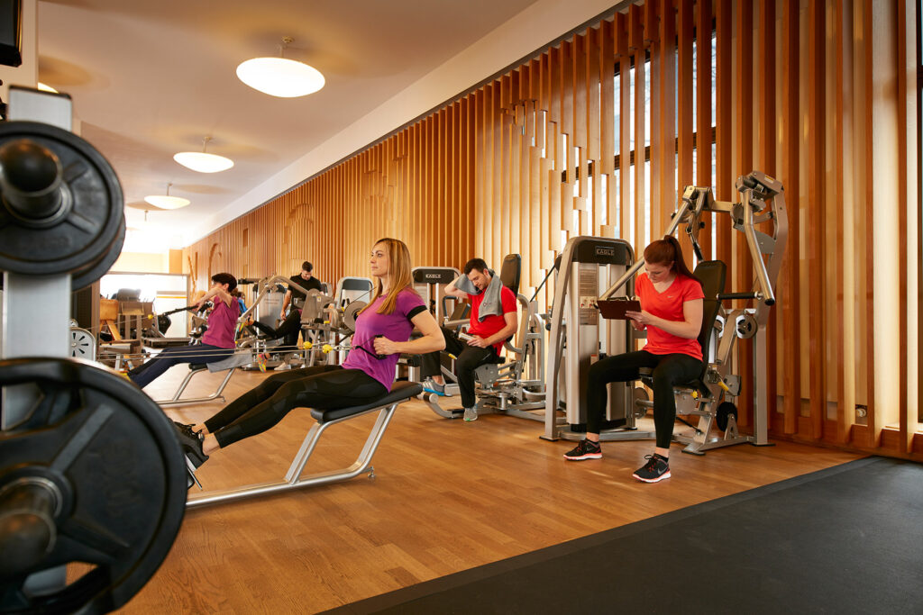 Menschen trainieren an verschiedenen Fitnessger&auml;ten in einem gut beleuchteten Fitnesscenter mit h&ouml;lzernen Wandpaneelen.