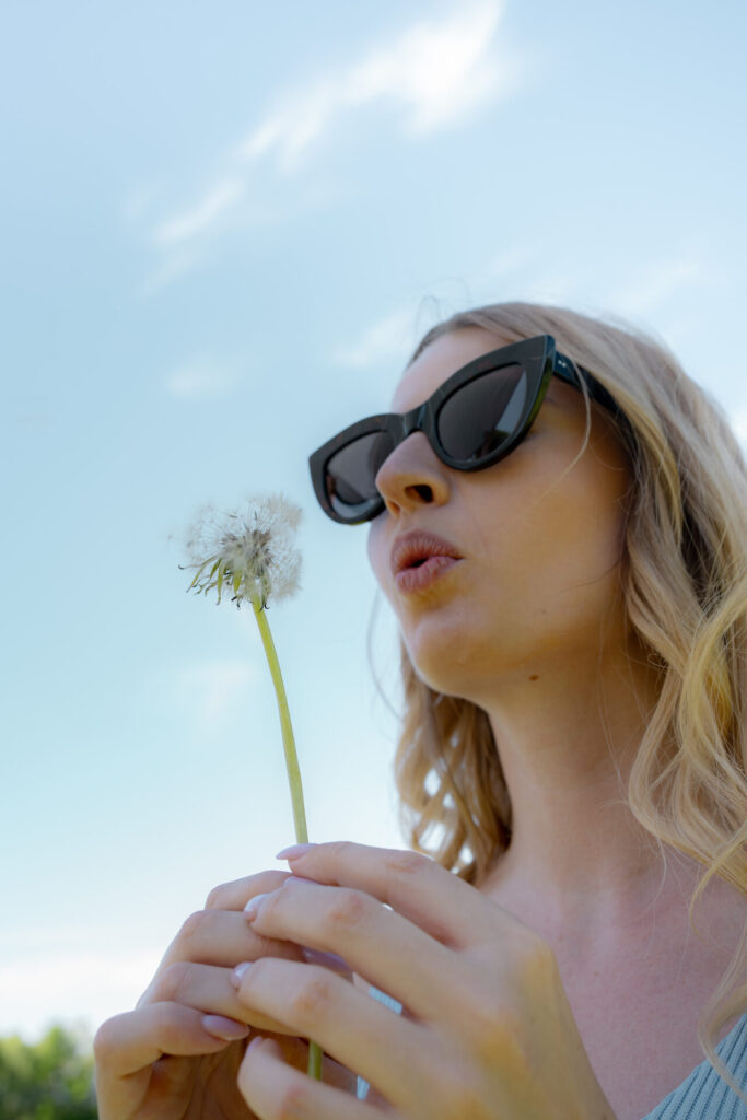 Frau mit Sonnenbrille pustet eine Pusteblume